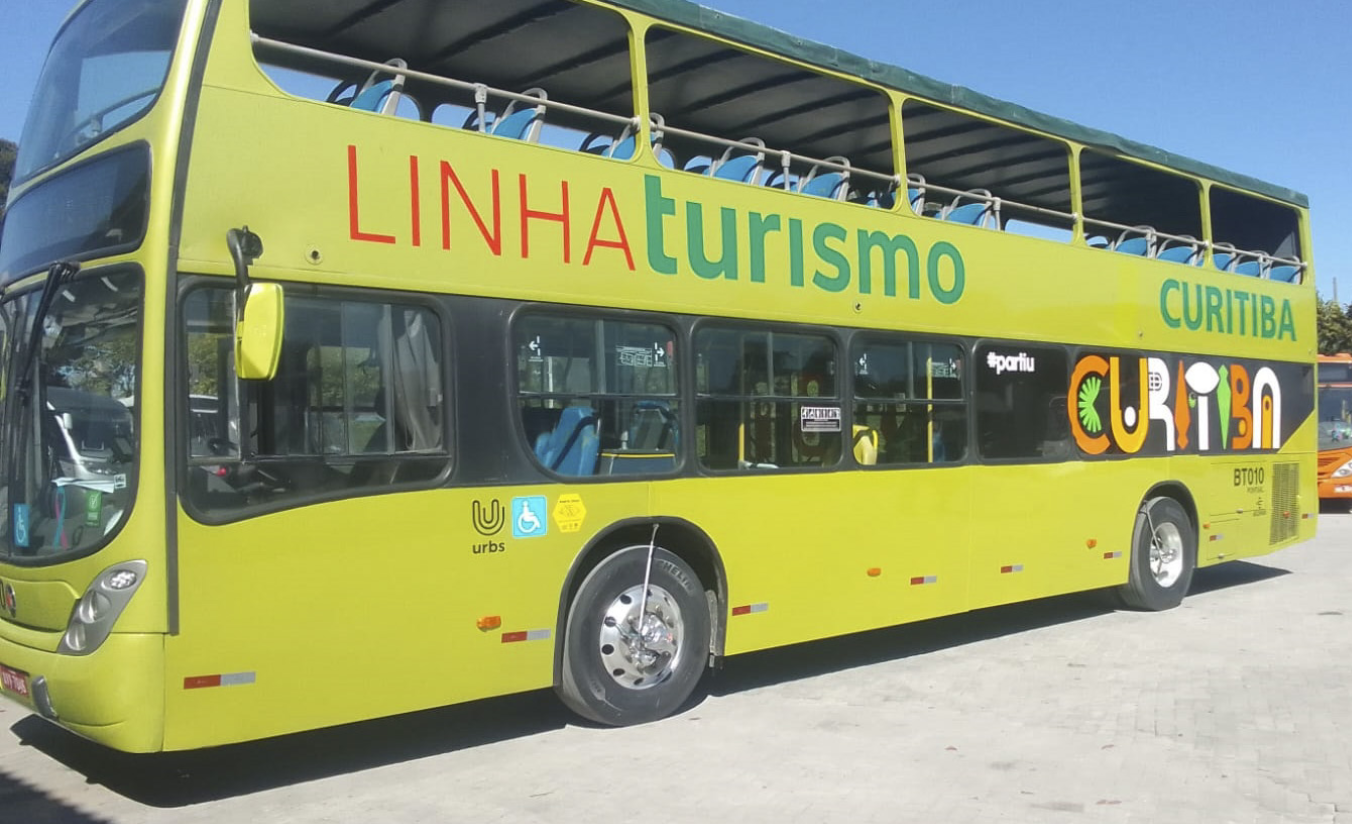 Ônibus Linha Turismo de Curitiba passando em frente a ponto turístico com araucárias ao fundo