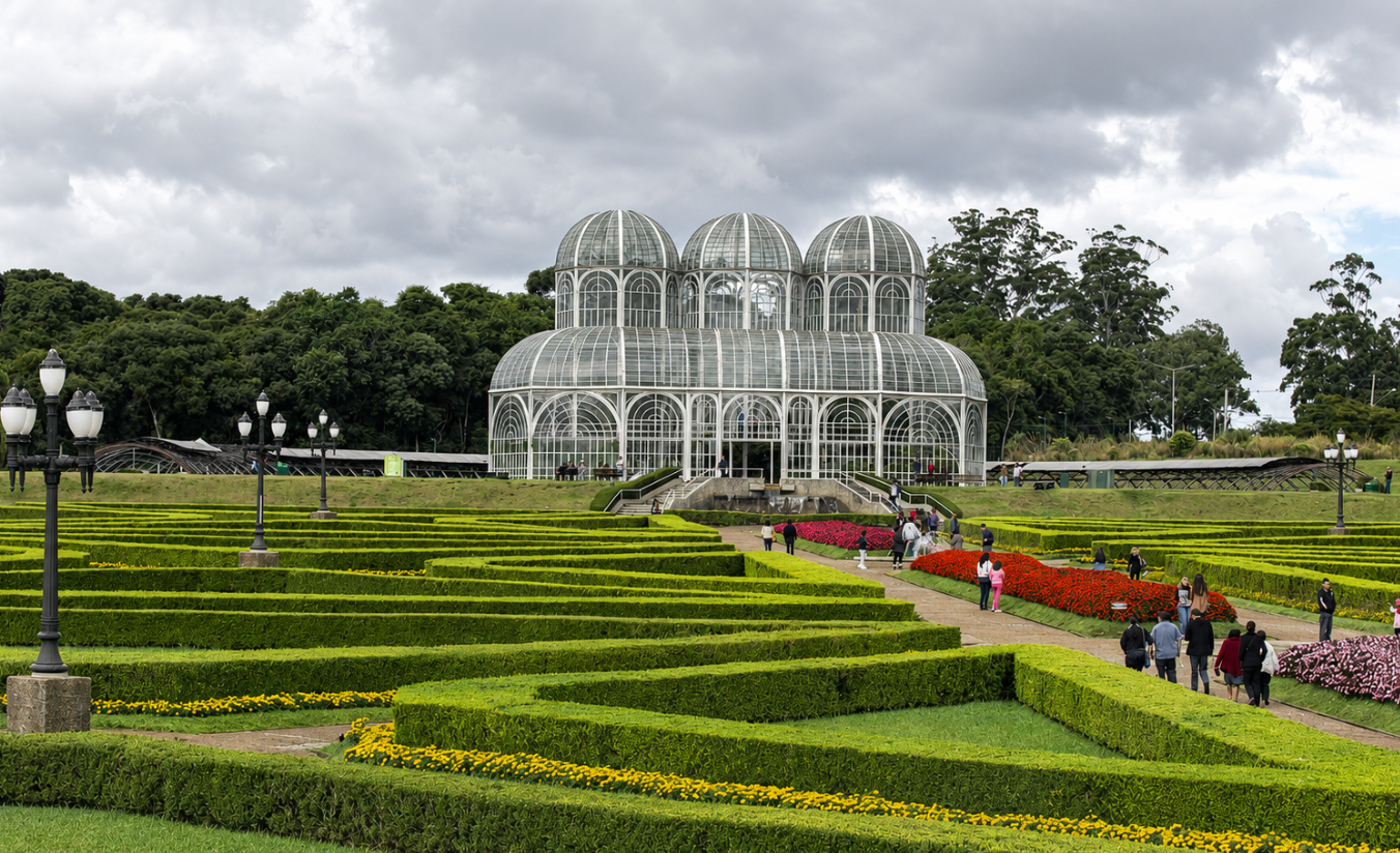 Jardim Botânico de Curitiba com a estufa de ferro e vidro ao fundo e os canteiros geométricos floridos em primeiro plano